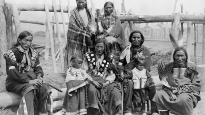 Lakota men, women, and children sit together in traditional clothing during the mid-19th century