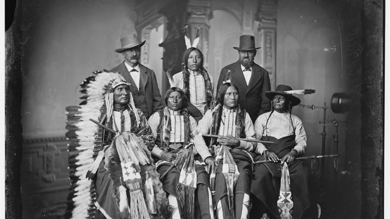 Sioux leaders in traditional dress sit for a formal photograph with two men in Western suits