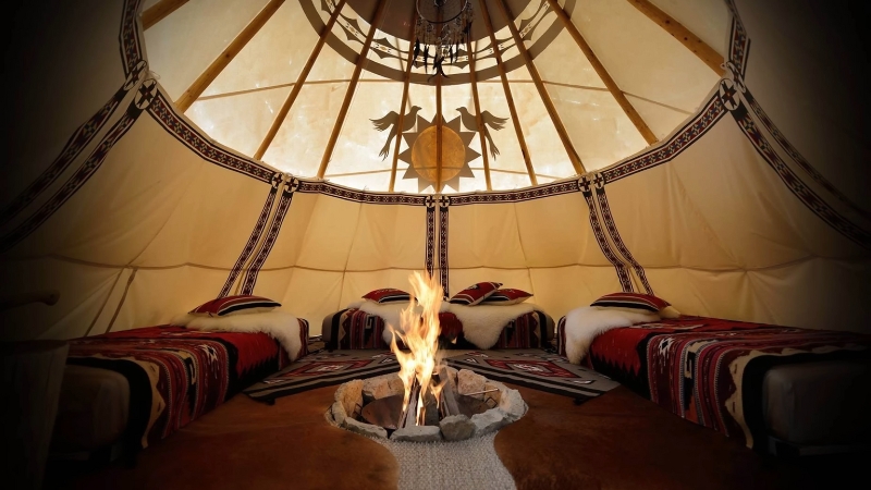 The inside of a Lakota tipi shows beds arranged in a circle around a central fire pit, with decorated walls and an open smoke hole above