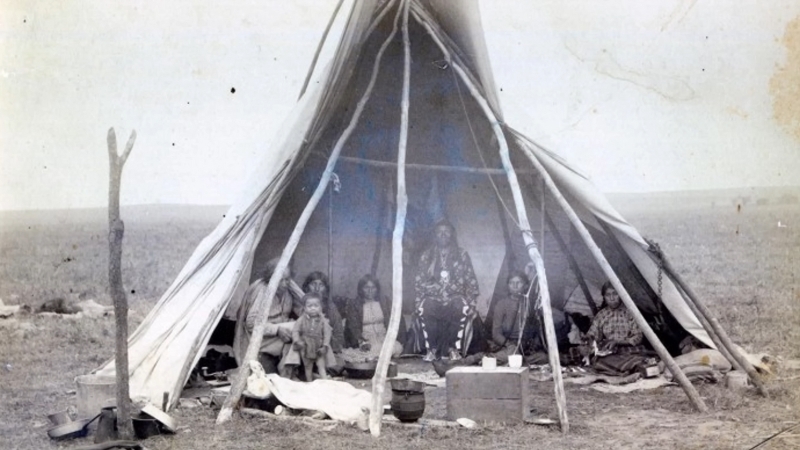 A Lakota family sits at the entrance of a partially open tipi on the plains
